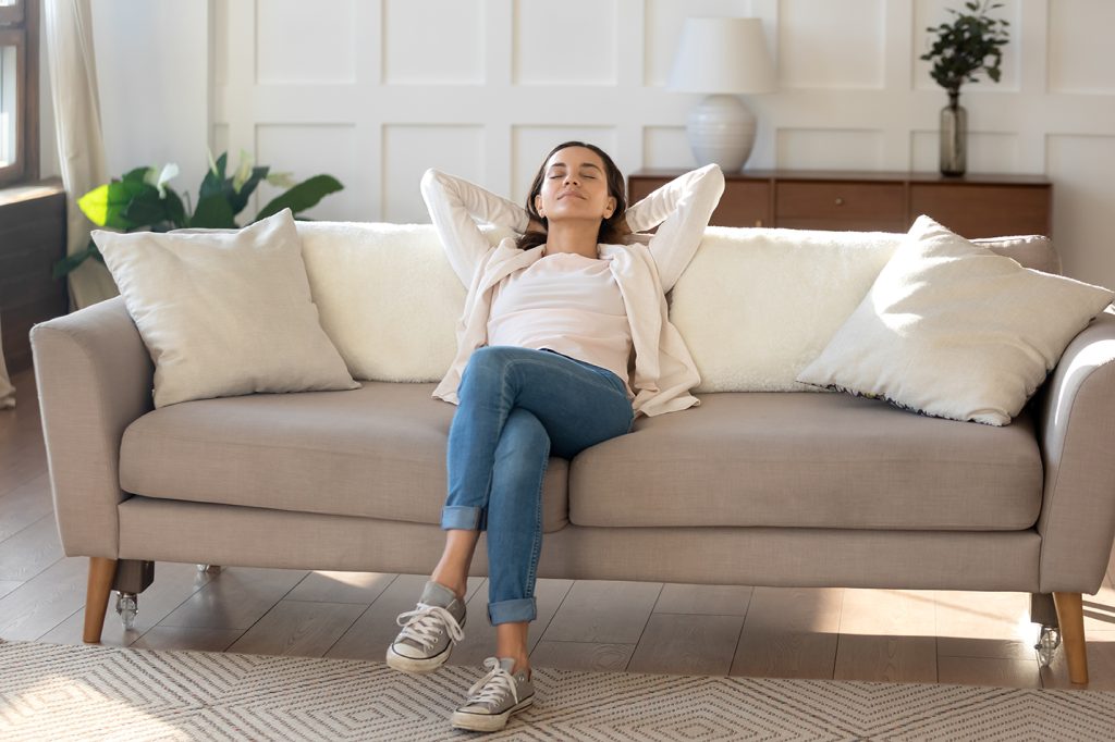 Calm and positive. Happy serene young female relaxing on couch at living room leaning back on pillows. Woman taking break of household chores, breathing fresh air, dreaming about spending weekend