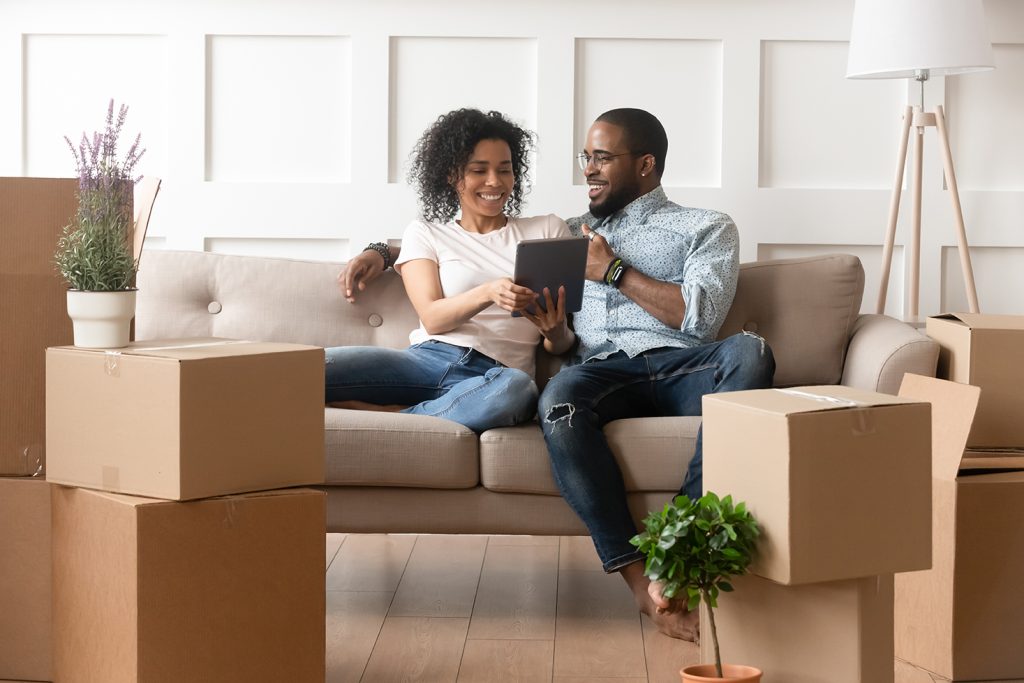 Happy african couple husband and wife choose house removal service or search renovating ideas sit on sofa with boxes, smiling black renters owners tenants use digital tablet on moving day in new home